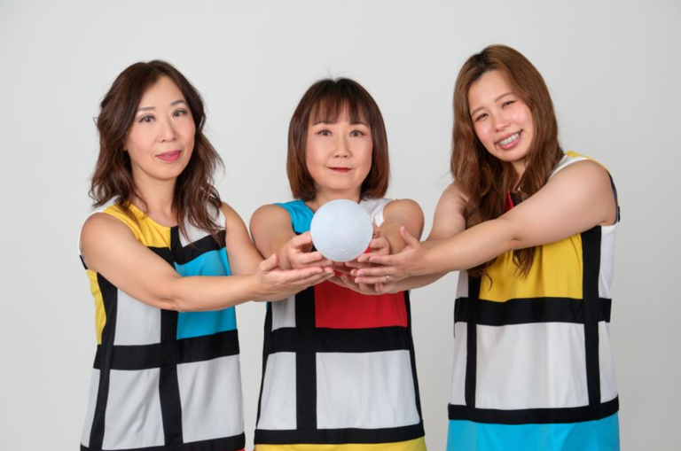 Pop-punk band Shonen Knife's three members wear matching Mondrian-style dresses and hold a white sphere together against a plain background.